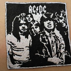 May include: Black and white embroidered patch featuring the band AC/DC. The patch has a rectangular shape with a black background and a white embroidered image of the band members. The band's name is embroidered at the top.