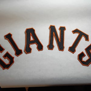 May include: Black and orange felt patches spelling out the word "GIANTS" in a curved arrangement. Each letter has a black base with an orange outline. The patches are on a white surface.