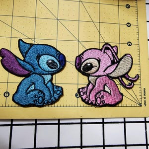 May include: Two embroidered patches of Stitch and Angel from the Disney movie Lilo & Stitch. Stitch is blue with purple accents and Angel is pink with white accents. Both are sitting with their arms crossed.