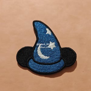May include: An embroidered patch shaped like a blue wizard hat with black trim and black mouse ears. The hat features a silver crescent moon and two silver stars. The text @Stitch_Kitten and Stitch kitten is embroidered in white across the center.