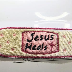 May include: Embroidered patch in the shape of a bandage. The patch has a light yellow background with pink accents and a pink border. The words "Jesus Heals" are embroidered in black with a red cross.