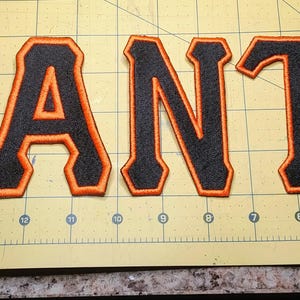 May include: Embroidered patches spelling "GIANTS" in black letters with orange trim. The patches are displayed on a yellow cutting mat with grid lines and measurement markings.