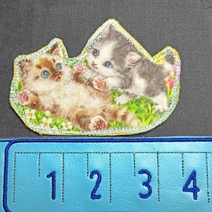 Op de afbeelding: Een stoffen patch met twee speelse kittens met blauwe ogen, tegen een achtergrond van groen gras en kleine bloemen. De patch heeft een gele rand en is ongeveer 7.6 cm breed.