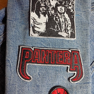 May include: Close-up of a denim jacket adorned with embroidered patches. The patches feature the AC/DC band logo, the word "PANTERA" in red and black, and a Spider-Man design. The jacket is a light blue wash.