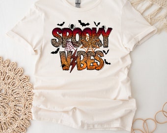 Op de afbeelding: Een wit T-shirt met een grafische print van de tekst "Spooky Vibes" in oranje, rood en zwart met een luipaardprint. De grafiek bevat ook vleermuizen, spinnen en spinnenwebben.
