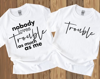 May include: Two white t-shirts with black text. One shirt says "nobody loves trouble as much as me" and the other shirt says "trouble".