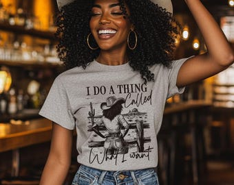 May include: A light gray t-shirt with the text "I DO A THING Called What I Want" and a graphic of a cowgirl. The cowgirl is wearing a hat and standing in front of a fence. The person in the image is wearing a cowboy hat and blue jeans.