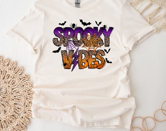 Op de afbeelding: Een wit T-shirt met een paarse, oranje en zwarte grafische print met de tekst "Spooky Vibes". De afbeelding bevat vleermuizen, spinnen en spinnenwebben.