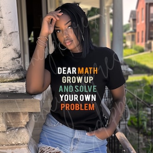 Könnte beinhalten: Schwarzes T-Shirt mit dem Text "DEAR MATH GROW UP AND SOLVE YOUR OWN PROBLEM" in Weiß, Orange und Grün. Das T-Shirt wird von einer Person mit dunklem Haar und blauen Jeansshorts getragen.