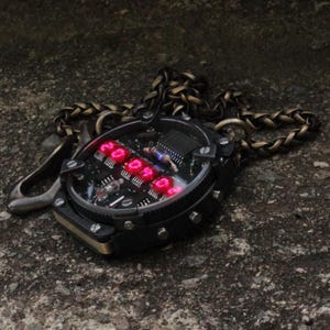 May include: A black and gold steampunk-style pendant watch with a red digital display showing the time. The watch has a clear face, a metal chain, and a clasp. The watch is set on a textured gray surface.