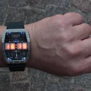 May include: A futuristic wristwatch with a rectangular silver case and a black strap. The watch face displays the time, 15:15, in bright orange digits. The watch has a complex circuit board design and is worn on a wrist.