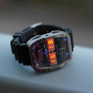 May include: A futuristic wristwatch with a silver rectangular case and a black rubber strap. The watch face displays red digital numbers and blue accent lights. The watch has a unique, tech-inspired design.