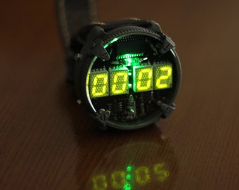 Könnte beinhalten: Eine schwarze und silberne Digitaluhr mit grüner Hintergrundbeleuchtung, die die Zeit 00:02 anzeigt. Die Uhr hat ein rundes Zifferblatt und ein dickes Armband.