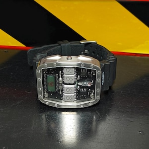 Puede incluir: Un reloj de pulsera con correa de caucho negra y caja rectangular de metal plateado. La esfera muestra un circuito impreso negro expuesto con componentes electrónicos visibles, incluyendo una pequeña pantalla digital y un tubo de vacío.