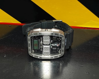 May include: A wristwatch with a black rubber strap and a rectangular silver-toned metal case. The watch face features an exposed black circuit board with visible electronic components, including a small digital display and a vacuum tube.