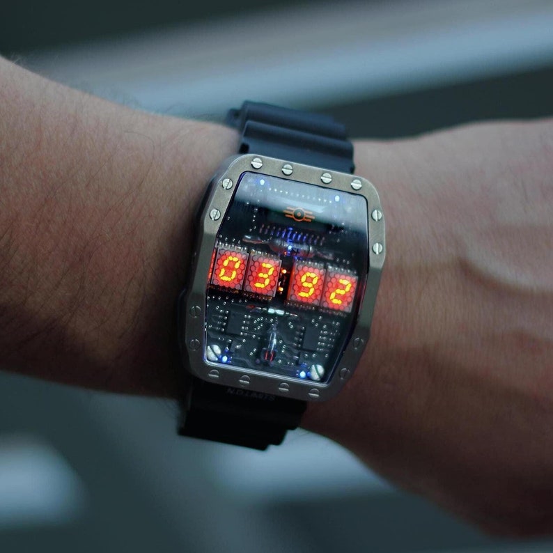 May include: A digital wristwatch with a rectangular stainless steel case and a black rubber strap. The watch face shows the time in orange numerals: 03:92. The watch has a circuit board design visible beneath the display. The strap is approximately 20 cm in length.