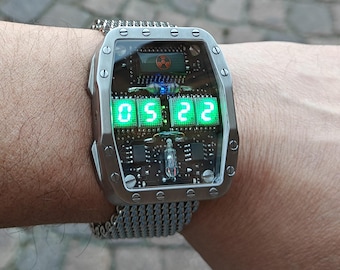 May include: A futuristic digital watch with a silver metal band and case. The watch face displays the time, 05:22, in bright green numerals. The watch has a unique design with visible internal components and a radiation symbol.