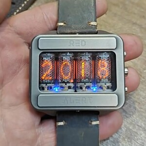 May include: A square, silver-toned wristwatch with a leather band. The watch face displays the time 2018 in glowing orange numerals within four glass tubes. The word "RED" is above the tubes, and "ALERT" is below.