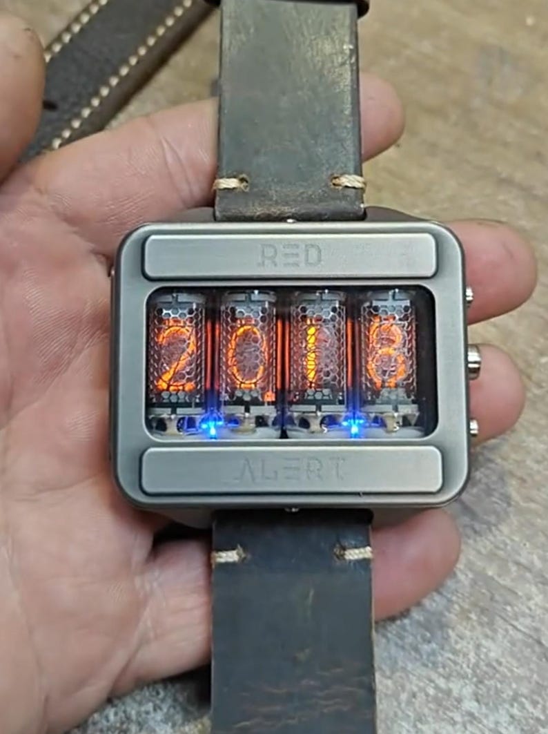 May include: A square, silver-toned wristwatch with a leather band. The watch face displays the time 2018 in glowing orange numerals within four glass tubes. The word "RED" is above the tubes, and "ALERT" is below.