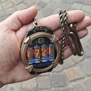Op de afbeelding: Een steampunk-stijl hangerhorloge met een bronskleurig frame en een carbon fiber achterkant. De wijzerplaat toont de tijd, 7:21, in oplichtende oranje cijfers in vier glazen buizen. Een bronzen ketting en sluiting zijn bevestigd.