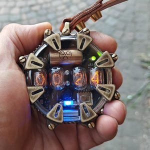 May include: A steampunk-style pendant necklace with a circular design. The pendant features glowing orange numerals, a radiation symbol, and a blue light. It is attached to a brown leather strap with brass hardware.