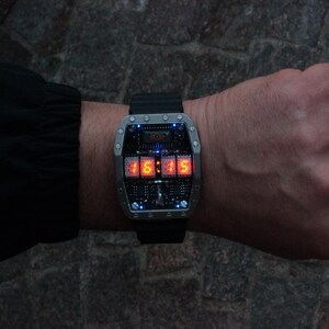 May include: A futuristic wristwatch with a black strap and a rectangular, silver-coloured face. The watch shows the time 15:15 in bright orange digital numbers. The face has a complex, circuit-board-like design with small blue lights.