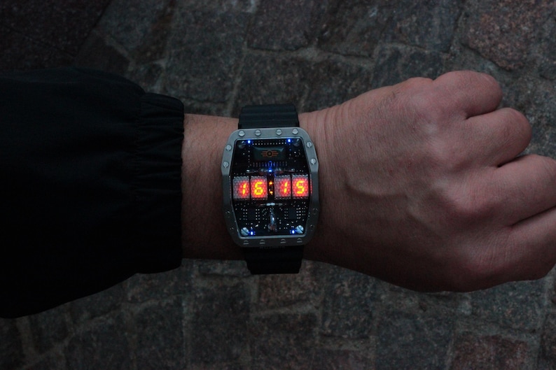 May include: A futuristic wristwatch with a black strap and a rectangular, silver-coloured face. The watch shows the time 15:15 in bright orange digital numbers. The face has a complex, circuit-board-like design with small blue lights.