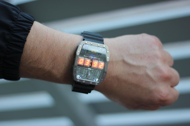 May include: A futuristic wristwatch with a silver case and a black rubber strap. The watch face displays the time in bright orange digital numbers. The watch is worn on a wrist.