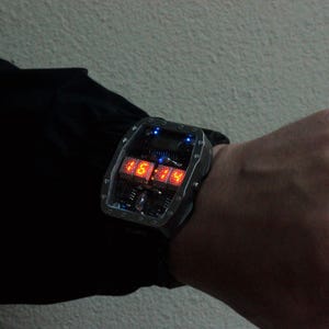 May include: A futuristic digital wristwatch with a rectangular, silver-coloured case. The watch displays the time in bright red numerals. The watch is worn on a wrist, with a black strap.