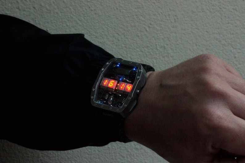 May include: A futuristic digital wristwatch with a rectangular, silver-coloured case. The watch displays the time in bright red numerals. The watch is worn on a wrist, with a black strap.