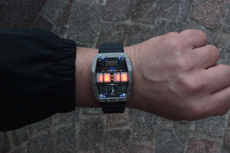 May include: A futuristic wristwatch with a rectangular silver case and a black strap. The watch face displays the time, 15:15, in bright orange digits. The watch has a complex circuit board design and is worn on a wrist.