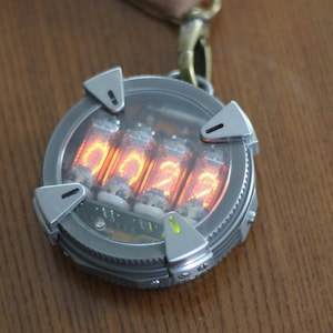 May include: A round, silver-colored digital clock with four glowing orange digits. The clock has a clear face and is attached to a leather strap with a metal clasp. The clock has a steampunk aesthetic.