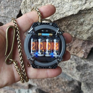 Pode incluir: Colar com pingente em estilo steampunk com um visor digital mostrando os números "20.03" em laranja brilhante. O pingente é envolto em uma moldura metálica com um detalhe em fibra de carbono e é suspenso por uma corrente dourada.