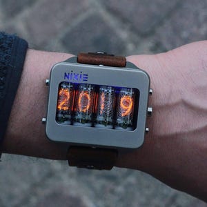 May include: A Nixie watch with a rectangular, gray metal case and a brown leather strap. The watch face displays the numbers "2019" in glowing orange digits. The word "Nixie" is printed above the numbers.