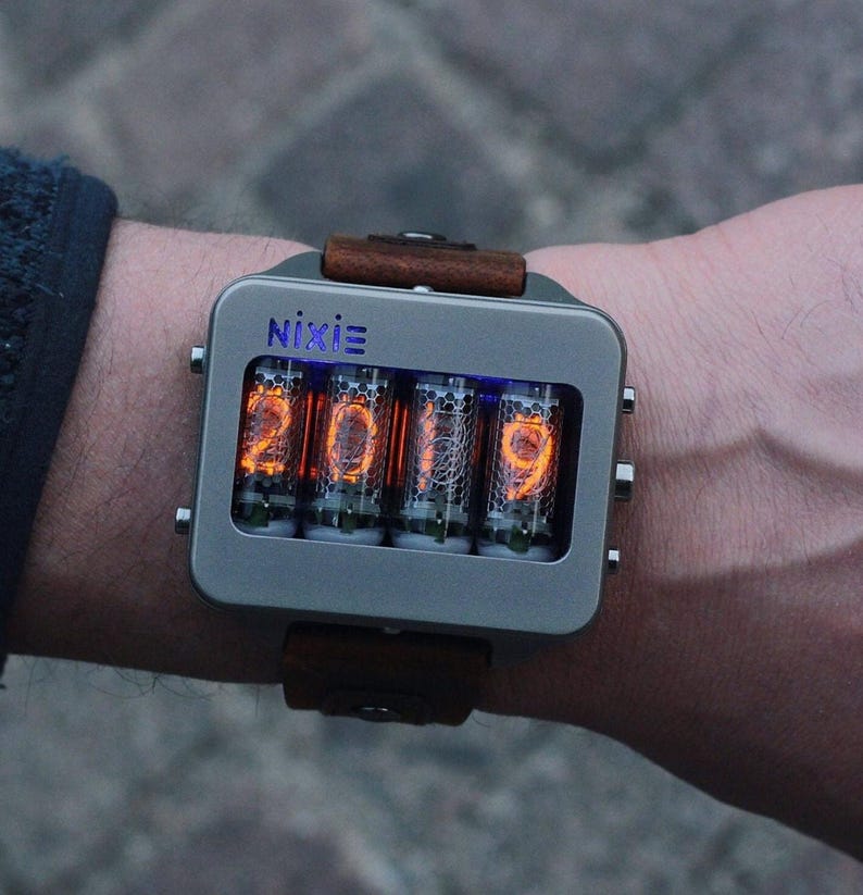 May include: A Nixie watch with a rectangular, gray metal case and a brown leather strap. The watch face displays the numbers "2019" in glowing orange digits. The word "Nixie" is printed above the numbers.
