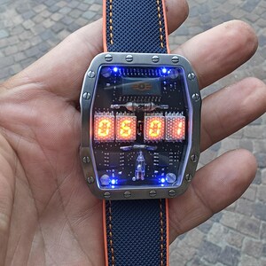 May include: A futuristic wristwatch with a rectangular stainless steel case and a blue and orange strap. The watch face displays the time in orange digital numerals against a black circuit board background. The watch is 4.5 cm wide.