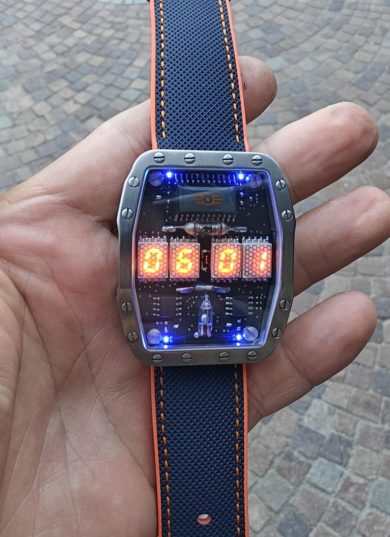 May include: A futuristic wristwatch with a rectangular stainless steel case and a blue and orange strap. The watch face displays the time in orange digital numerals against a black circuit board background. The watch is 4.5 cm wide.