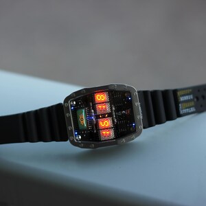 May include: A digital wristwatch with a black rubber strap. The watch face displays orange digital numbers and a green graphic. The watch has a silver-coloured metal casing. The watch is on a light blue surface.