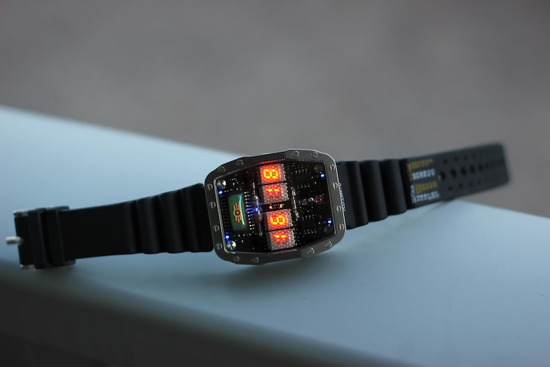 May include: A digital wristwatch with a black rubber strap. The watch face displays orange digital numbers and a green graphic. The watch has a silver-coloured metal casing. The watch is on a light blue surface.