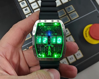 May include: A futuristic wristwatch with a black band and a rectangular, silver-colored face. The watch displays the time as 12:10 in bright green digital numbers. The face has a circuit board design with a green glow.