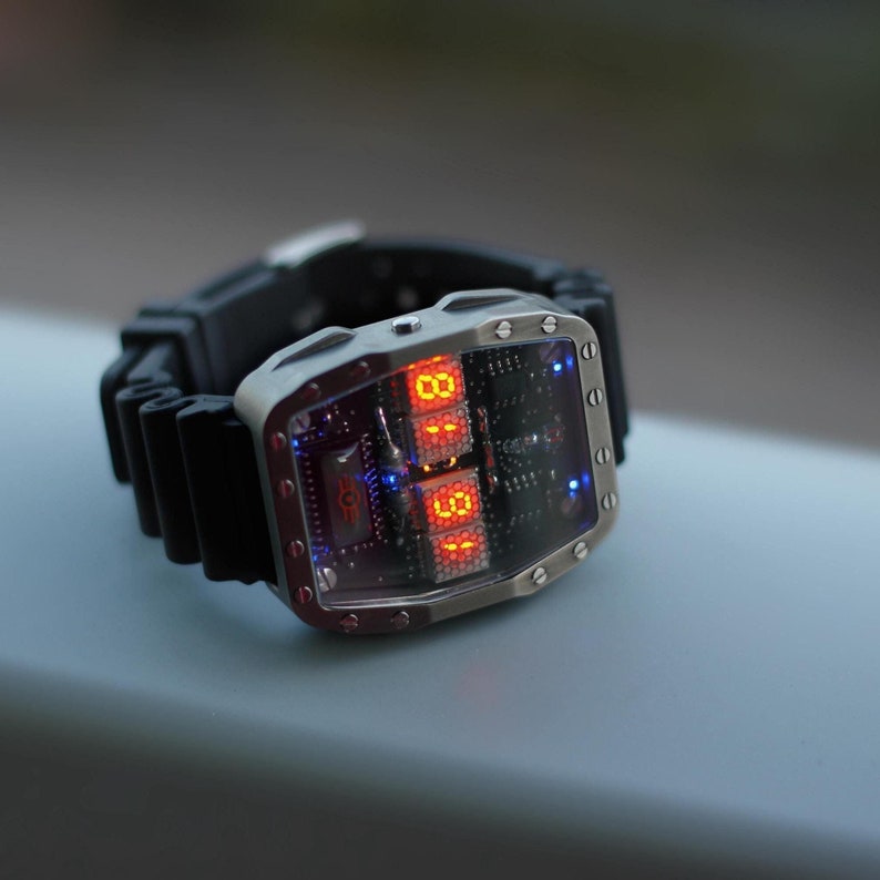 May include: A futuristic wristwatch with a silver rectangular case and a black rubber strap. The watch face displays red digital numbers and blue accent lights. The watch has a unique, tech-inspired design.