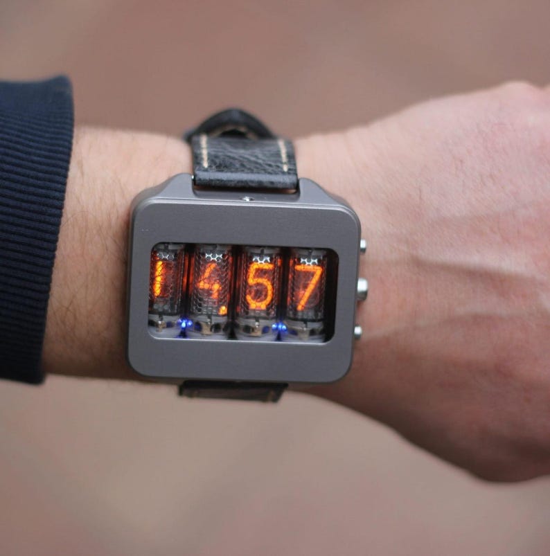 Può includere: Un orologio da polso digitale grigio con cinturino in pelle nera mostra l'ora 14:57 in numeri arancioni. L'orologio ha un quadrante rettangolare con quattro tubi illuminati. L'orologio è indossato al polso.