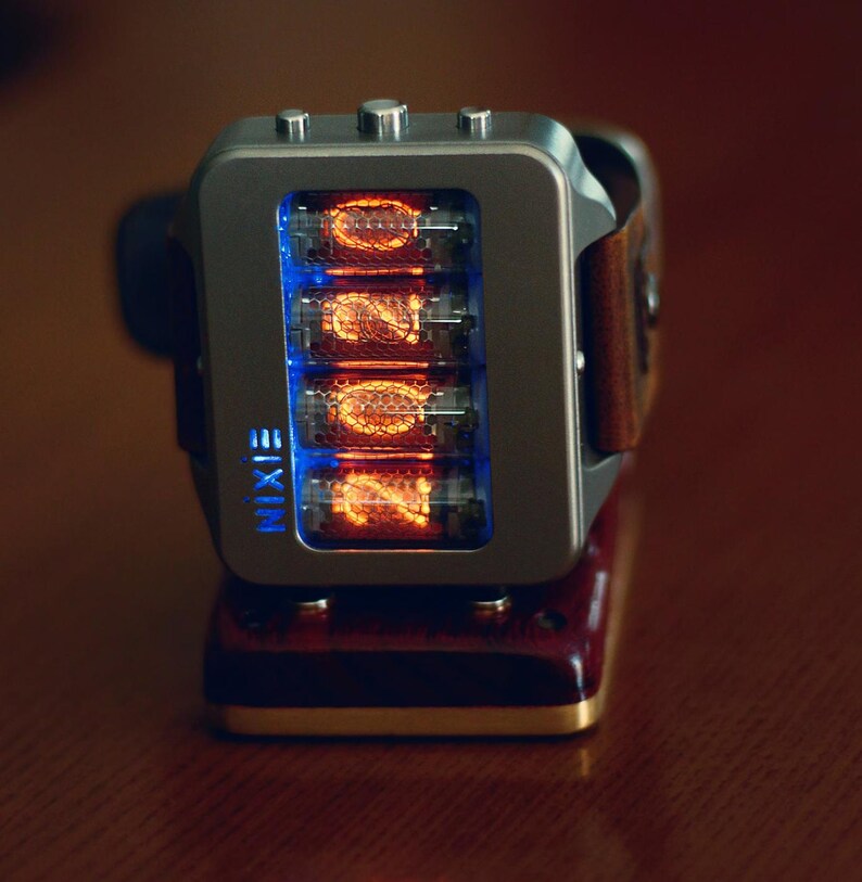 May include: A silver Nixie tube watch with a brown leather strap. The watch face displays four glowing orange tubes with blue backlighting. The word "NIXIE" is printed in blue on the watch face.
