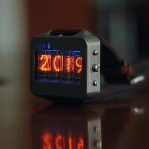 May include: A Nixie watch displaying the year 2019 in bright orange digits. The watch has a rectangular, silver-colored case with three buttons on the side and a dark strap. The word "Nixie" is printed on the top of the watch face.