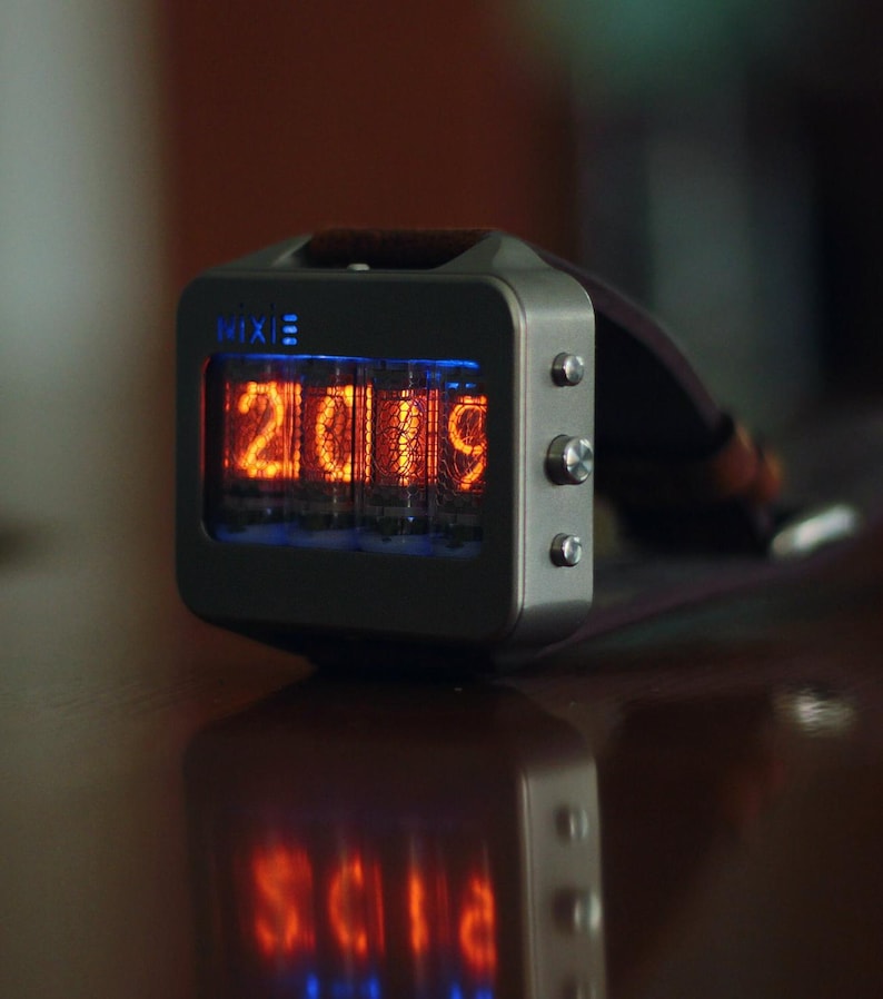 May include: A Nixie watch displaying the year 2019 in bright orange digits. The watch has a rectangular, silver-colored case with three buttons on the side and a dark strap. The word "Nixie" is printed on the top of the watch face.