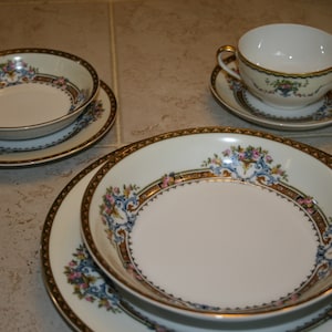 May include: A set of white porcelain dishes with a floral design in pink, blue, and gold. The set includes bowls, plates, and a handled cup and saucer. The rims are trimmed with a gold and brown border.