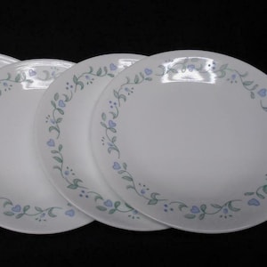 May include: A set of white ceramic plates with a delicate floral design. Each plate features a border of green vines, blue hearts, and small blue dots. The plates are arranged in a slightly overlapping pattern.