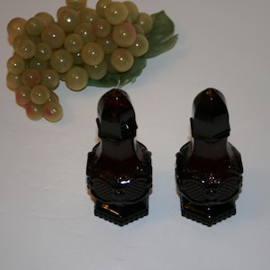 May include: Two dark red glass perfume bottles shaped like knights, with detailed armor and helmets. The bottles are next to a bunch of green grapes on a white surface. The bottles have a detailed design.