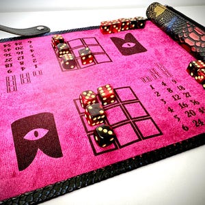 May include: A pink felt dice rolling mat with a black leather border and a black and white eye design. The mat has a grid for rolling dice and numbers printed on the side. There are red and black dice on the mat.