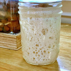 May include: A glass jar with a silver lid filled with a white, bubbly mixture. The jar is sitting on a wooden surface.